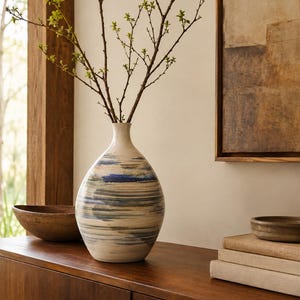 May include: A ceramic vase with a rounded body and a narrow neck, featuring horizontal brushstrokes in shades of blue, brown, and cream. The vase contains thin, bare branches with small green leaves extending from the top opening.