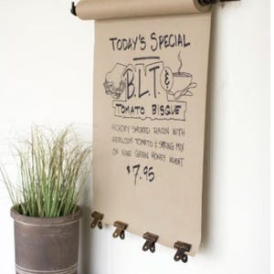 Könnte beinhalten: Eine braune Papierrolle mit einem Metallhalter, der an einer weißen Wand befestigt ist. Das Papier zeigt eine handgeschriebene Speisekarte für das Tagesgericht: BLT & Tomatensuppe für 7,95 €.
