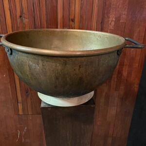 May include: A large, round, copper pot with two handles. The pot is sitting on a wooden pedestal against a wood panel background.