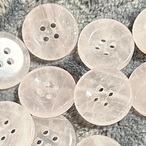 Może przedstawiać: Dziesięć okrągłych, przezroczystych różowych guzików z czterema otworami każdy. Guziki są wykonane z materiału przypominającego kwarc lub szkło.