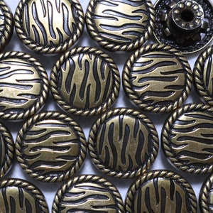 May include: A collection of round, gold-toned metal buttons with a black and gold zebra stripe pattern. The buttons have a rope-like border.
