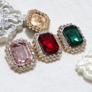 以下が含まれることがあります： 金色の枠に囲まれたカラフルなガラス製の宝石が4つ。宝石はピンク、赤、緑、金で、透明なラインストーンで囲まれています。