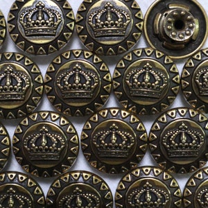 Pode incluir: Close-up de vários botões antigos de cor latão. Cada botão apresenta um design de coroa detalhado no centro, rodeado por uma borda decorativa. Um botão tem uma parte traseira diferente, sugerindo um fecho de pressão. Os botões estão próximos.