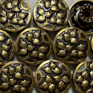 Op de afbeelding: Een verzameling van 14 ronde, goudkleurige knopen met een zwarte achtergrond en een reliëfdesign van bladeren. De knopen hebben een glanzende afwerking en een decoratieve rand.