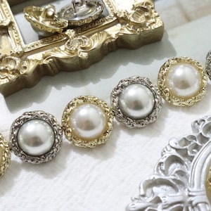 Könnte beinhalten: Nahaufnahme einer Reihe von perlmuttartigen Knöpfen mit goldenen und silbernen Einstellungen. Die Knöpfe sind in einer Linie auf einer weißen Oberfläche angeordnet.