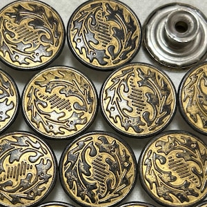 Könnte beinhalten: Eine Sammlung von 14 goldenen und schwarzen Knöpfen mit einem dekorativen Blattdesign. Die Knöpfe sind rund und haben ein erhabenes Design. Es gibt auch einen einzelnen silbernen Knopf mit einem Loch in der Mitte.