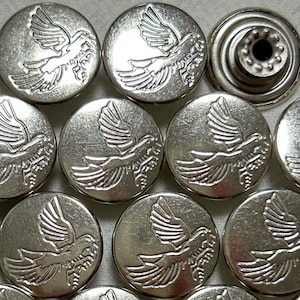 Könnte beinhalten: Ein Set mit 13 silberfarbenen Metallknöpfen mit einem Taubenmotiv. Die Knöpfe sind rund und haben ein erhabenes Taubenmotiv in der Mitte. Die Knöpfe sind perfekt, um jedem Kleidungsstück einen Hauch von Eleganz zu verleihen.