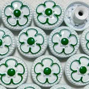 May include: A collection of 12 white and green floral buttons with a raised design. The buttons have a green center and a white outer ring with a green floral design.