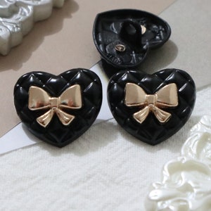May include: Two black heart-shaped buttons with a gold bow design. The buttons have a quilted texture.