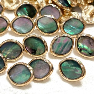 Może przedstawiać: Zbiór owalnych, iryzujących zielono-fioletowych guzików muszlowych z pozłacanymi metalowymi obwódkami. Guziki mają lekko nieregularny kształt i fakturowaną powierzchnię.