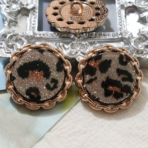 May include: Two gold-toned buttons with a leopard print design and glitter accents. The buttons have a decorative, rope-like border.