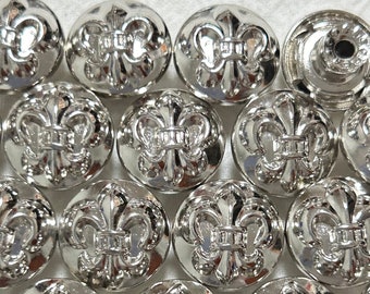 Puede incluir: Una colección de botones de metal plateados con un diseño de flor de lis. Los botones son redondos y tienen un diseño en relieve.