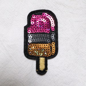 May include: A colourful sequined ice cream cone patch with pink, silver, and gold sequins. The patch is perfect for adding a fun and whimsical touch to clothing, bags, or other accessories.