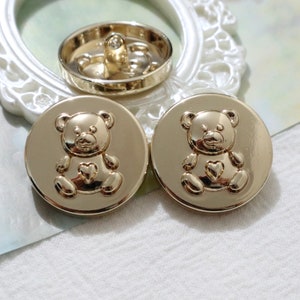 May include: Two gold-tone metal buttons featuring a teddy bear design. Each button has a raised heart on the teddy bear's chest.