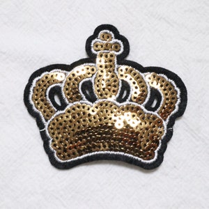 Op de afbeelding: Een gouden pailletten kroon patch met zwarte bies. De kroon is een populair motief voor kleding en accessoires.