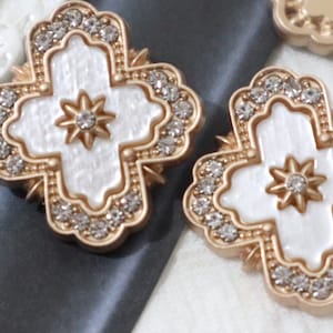 Könnte beinhalten: Zwei verzierte, goldfarbene kreuzförmige Verzierungen mit weißer Mitte und Sternmuster. Die Kreuze sind mit kleinen, klaren Kristallen an den Rändern besetzt. Der Hintergrund ist eine Mischung aus Grau und Weiß.