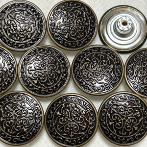 Może przedstawiać: Zestaw dziesięciu antycznych, złotych metalowych guzików z ozdobnym wzorem w kolorze czarnym i złotym. Guziki mają wypukły, okrągły kształt z ozdobnym wzorem.