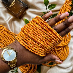 May include: A stack of orange and yellow beaded bracelets. A gold watch with a light blue face is on the wrist. A bottle of cologne and green leaves are in the background.