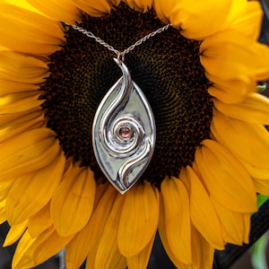 May include: A silver pendant necklace with a teardrop-shaped design, featuring a central spiral and a small pink gemstone. The pendant hangs on a delicate silver chain, set against a bright yellow sunflower.