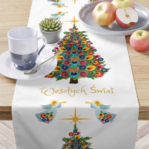 May include: White table runner featuring a colourful folk art Christmas tree design. The tree is adorned with red, yellow, blue, green and pink flowers. The runner also includes two angels with trumpets and the text "Wesołych Świąt" in gold lettering.