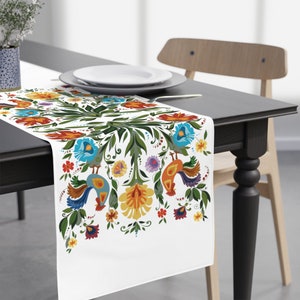 Puede incluir: Caminero de mesa blanco con un diseño floral y de gallo colorido. El diseño incluye hojas verdes, flores rojas, azules, amarillas y naranjas, y dos gallos con plumaje colorido.