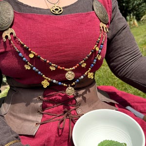 Könnte beinhalten: Ein dunkelrotes Leinen-Schürzenkleid über einem dunkelbraunen Langarmshirt, mit zwei großen ovalen Bronzebroschen, die durch zweireihige Perlenketten mit kleinen goldfarbenen Anhängern verbunden sind, sowie einem braunen Schnürkorsett.