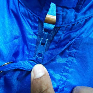 May include: Close-up of a blue zipper on a blue jacket. The zipper is metal with a blue slider.