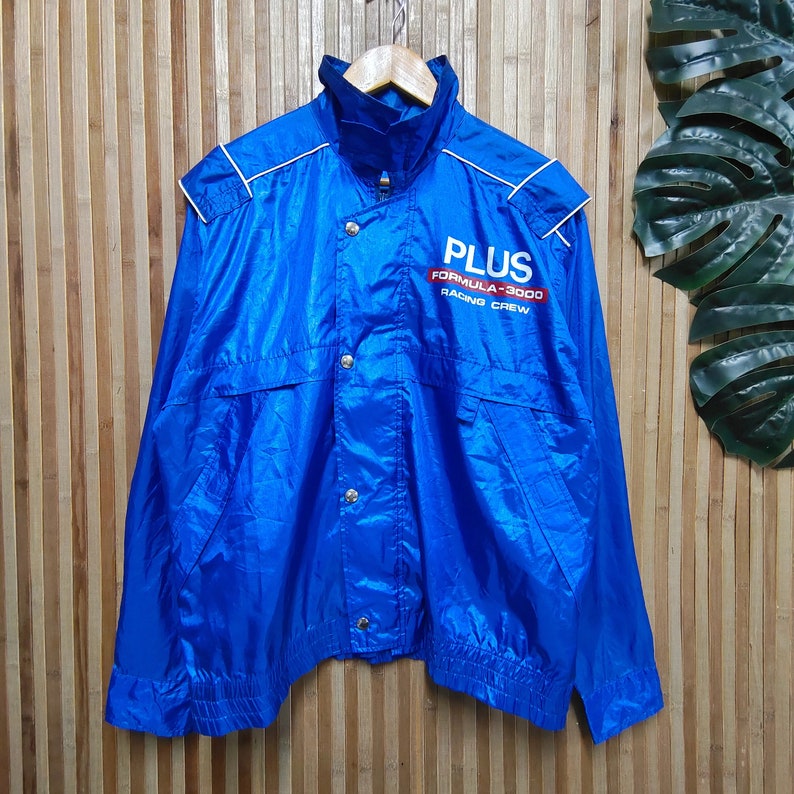 May include: A blue nylon jacket with a white zipper and white piping. The jacket has a logo that says 'PLUS FORMULA-3000 RACING CREW' in red and white lettering.