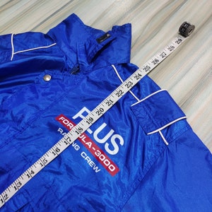 May include: A blue nylon jacket with white piping. The jacket has a zipper closure and a white drawstring hood. The jacket has a white and red logo that says 'Formula-3000 Racing Crew'.