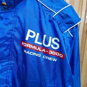 May include: A blue nylon jacket with white piping. The jacket has a white zipper and a white collar. The jacket has a red and white logo that says 'PLUS FORMULA-3000 RACING CREW'.