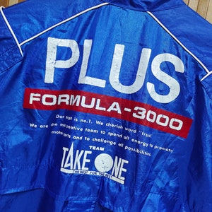 May include: A blue jacket with white piping and the words 'PLUS FORMULA-3000' in white and red lettering. Below the text is a white circle with the words 'TEAM TAKE ONE' and 'THE BEST FOR THE BEST' in black lettering.