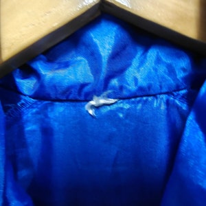 May include: A close-up of the inside of a blue jacket collar. The collar is made of a smooth, shiny fabric. A small white tag is attached to the collar.