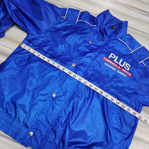 May include: A blue nylon jacket with a white zipper and white piping. The jacket has a  'PLUS FORMULA-3000 RACING CREW' logo on the front. A measuring tape is laid across the jacket.
