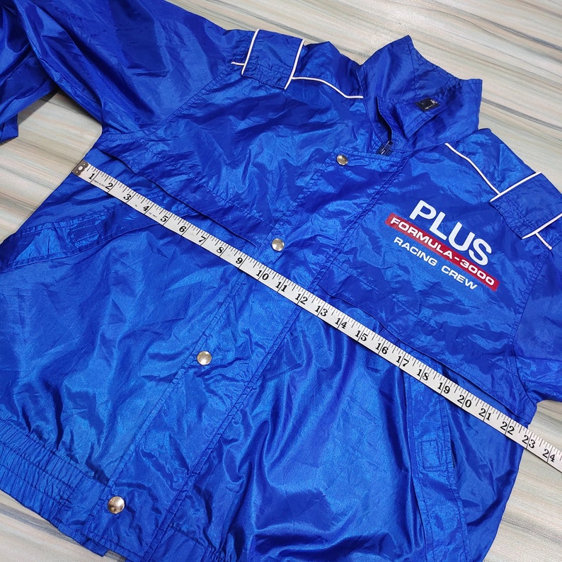 May include: A blue nylon jacket with a white zipper and white piping. The jacket has a  'PLUS FORMULA-3000 RACING CREW' logo on the front. A measuring tape is laid across the jacket.