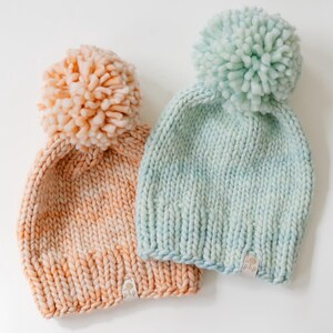 May include: Two chunky knit beanies, one in peach and one in light mint green, each topped with a large matching pom-pom. Both hats feature a thick ribbed brim and a small rectangular white fabric tag with the text "plp" at the bottom edge.