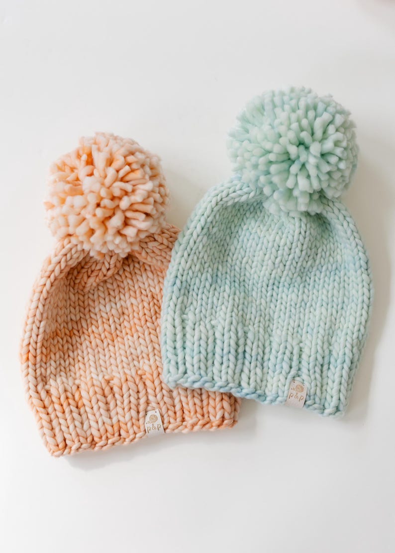 May include: Two chunky knit beanies, one in peach and one in light mint green, each topped with a large matching pom-pom. Both hats feature a thick ribbed brim and a small rectangular white fabric tag with the text "plp" at the bottom edge.