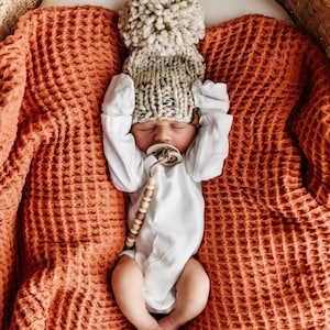 May include: A newborn baby wearing a white long-sleeve bodysuit and a chunky knit beige beanie with a large pom-pom. The baby has a beige dummy with a wooden bead clip attached, resting on a textured rust-coloured waffle-knit blanket.