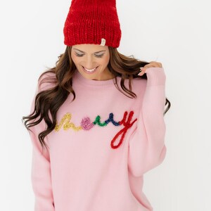 May include: A light pink long-sleeve jumper featuring the word merry embroidered in multi-coloured, textured yarn letters in yellow, pink, green, and red. The jumper is paired with a bright red knit beanie topped with a large matching pom-pom.