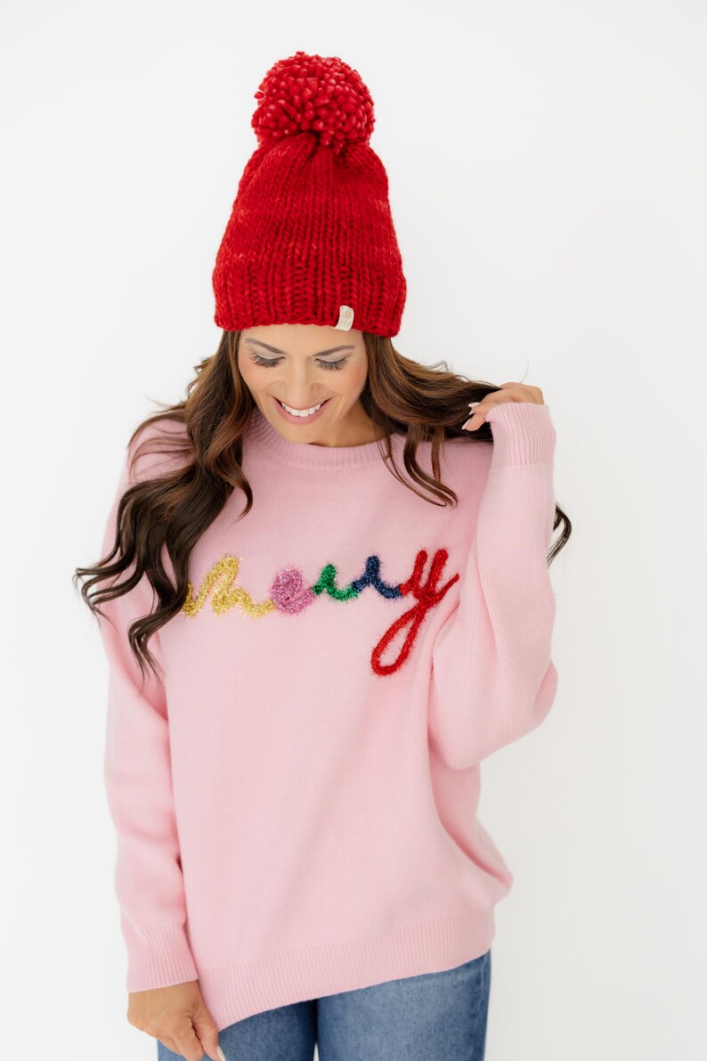 May include: A light pink long-sleeve jumper featuring the word merry embroidered in multi-coloured, textured yarn letters in yellow, pink, green, and red. The jumper is paired with a bright red knit beanie topped with a large matching pom-pom.
