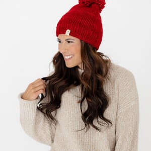 May include: A bright red knit beanie featuring a large, matching red pom-pom on top. The hat is crafted from a chunky yarn with a visible ribbed texture and includes a small, rectangular beige brand tag attached to the lower edge.