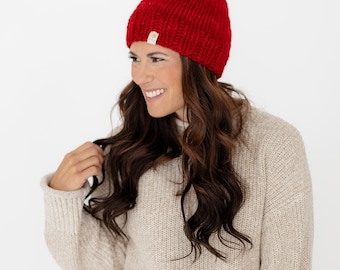 May include: A bright red knit beanie featuring a large, matching red pom-pom on top. The hat is crafted from a chunky yarn with a visible ribbed texture and includes a small, rectangular beige brand tag attached to the lower edge.