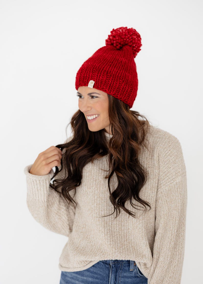 May include: A bright red knit beanie featuring a large, matching red pom-pom on top. The hat is crafted from a chunky yarn with a visible ribbed texture and includes a small, rectangular beige brand tag attached to the lower edge.