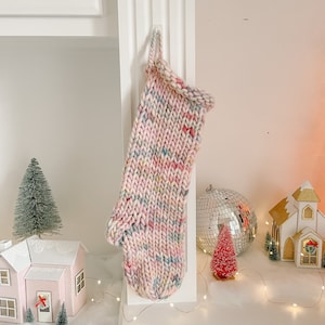 Puede incluir: Una media de Navidad de punto rosa y blanco con motas de colores cuelga de un gancho blanco. La media está decorada con un pompón blanco en la parte superior. La media está colgada en un estante blanco con una pequeña casa rosa, un pequeño árbol verde y una bola de discoteca plateada.