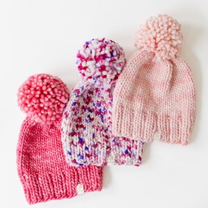 May include: Three chunky knit beanies with large pom-poms on top. The left beanie is solid dark pink, the centre beanie features a multi-coloured speckled pattern of pink, purple, and white, and the right beanie is a soft, pale pink.