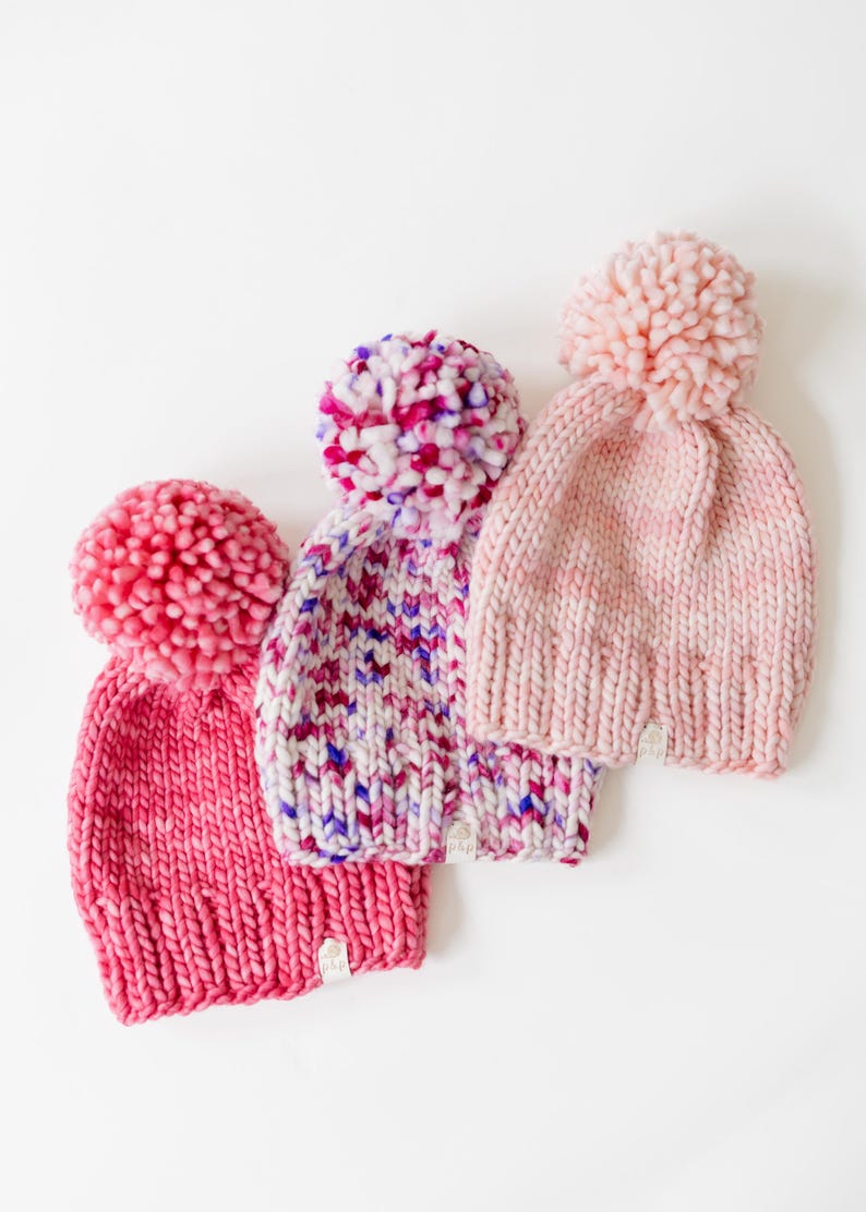 May include: Three chunky knit beanies with large pom-poms on top. The left beanie is solid dark pink, the centre beanie features a multi-coloured speckled pattern of pink, purple, and white, and the right beanie is a soft, pale pink.