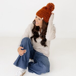 May include: A chunky knit beanie in a deep burnt orange colour featuring a large matching pom-pom on top and a small rectangular white fabric tag on the folded brim. The hat is made of thick yarn with a visible textured stitch pattern.