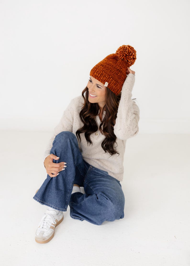 May include: A chunky knit beanie in a deep burnt orange colour featuring a large matching pom-pom on top and a small rectangular white fabric tag on the folded brim. The hat is made of thick yarn with a visible textured stitch pattern.
