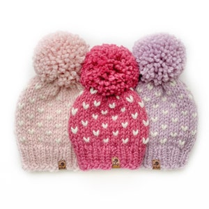 May include: Three knitted hats with pom-poms in pink, lavender, and red with white heart patterns. Each hat has a brown tag with the text "p&p" on it.