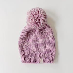 May include: A chunky knit beanie in a variegated light purple and white yarn. The hat features a large, matching pom-pom on top and a ribbed brim. A small, rectangular fabric tag with the text p&p is sewn onto the lower edge of the beanie.