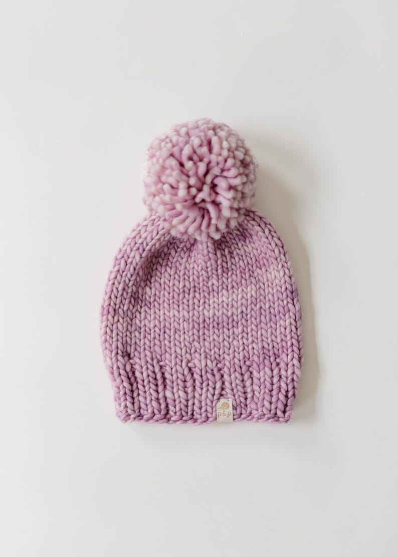 May include: A chunky knit beanie in a variegated light purple and white yarn. The hat features a large, matching pom-pom on top and a ribbed brim. A small, rectangular fabric tag with the text p&p is sewn onto the lower edge of the beanie.
